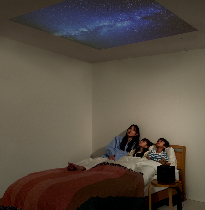 子ども2人と母親がベッドの上で天井に映し出された映像を見ています